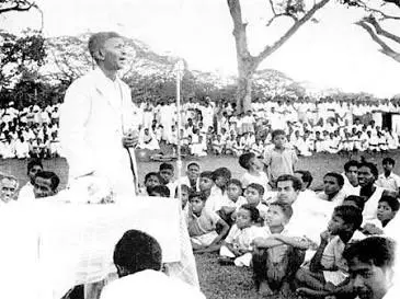 Amirthalingam en costume clair parle au micro devant une foule assise en extérieur.