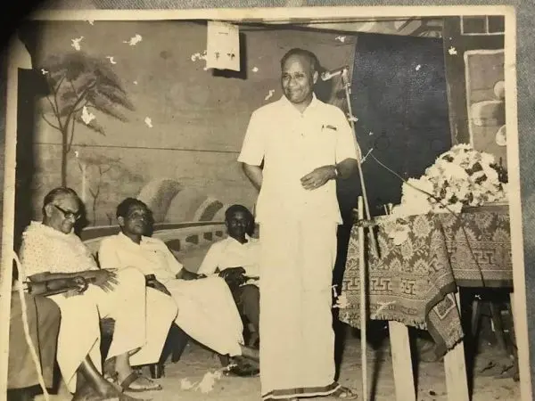 Amirthalingam en chemise et veshti blancs, au micro, avec des hommes assis à côté.