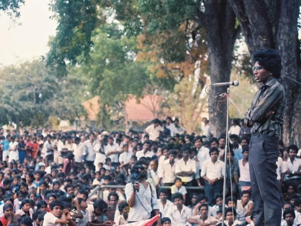 Thileepan au micro, bras croisés, devant une foule assise sous les arbres.