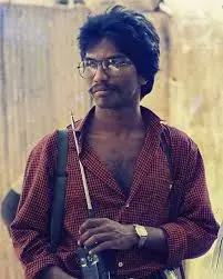 Portrait de Thileepan, lunettes et chemise à carreaux, un fusil sur l'épaule.